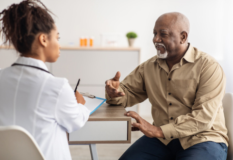 Older Black man in button-down shirt speaking with a young, Black, female doctor who is making eye contact and holding a pen and clip board