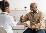 An older Black man talking to a younger Black doctor who is listening with a pad to take notes